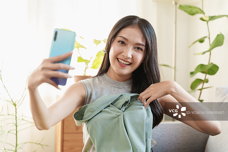 年轻漂亮亚洲女人在家试衣服时用手机自拍图片素材