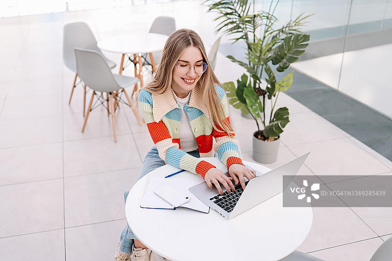 面带微笑的年轻女大学生戴着眼镜，身穿亮色毛衣，在咖啡馆的公共场所坐在桌旁，使用手机和笔记本电脑在线学习和工作。在线教育。现代办公室图片素材