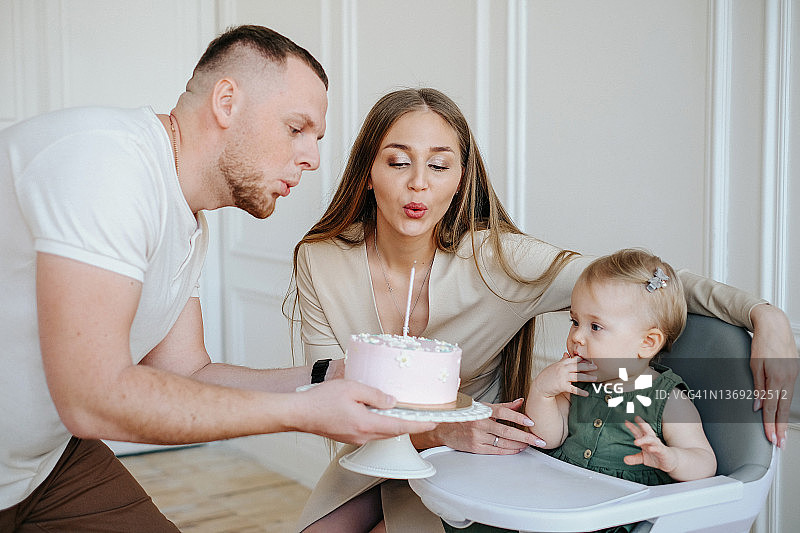 小女孩一周岁生日，幸福的父母给孩子送蛋糕，蛋糕上有一根蜡烛，美丽现代家庭的节日图片素材