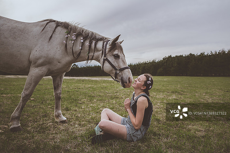 青少年女孩和一匹编着辫子、戴着鲜花的灰色马坐在一起图片素材