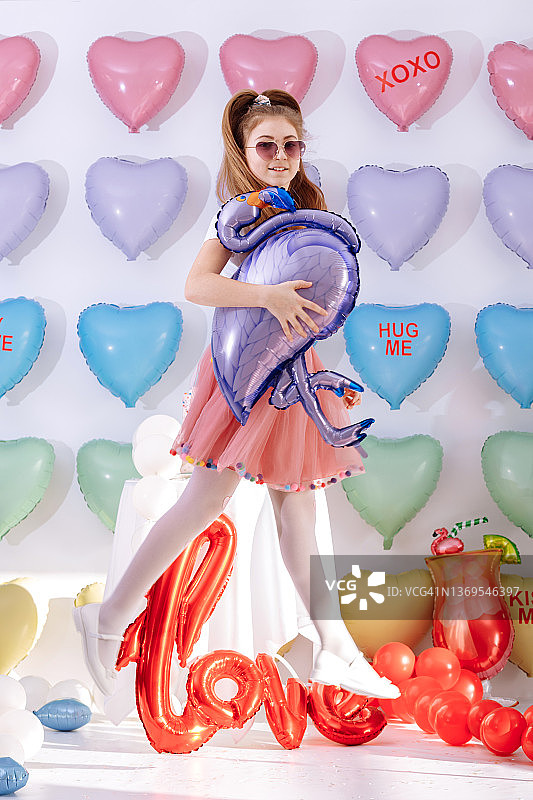 戴着太阳镜的可爱小女孩手拿火烈鸟气球，背景是彩色心形气球。微笑，有趣的 Valentine s Day 生日派对图片素材