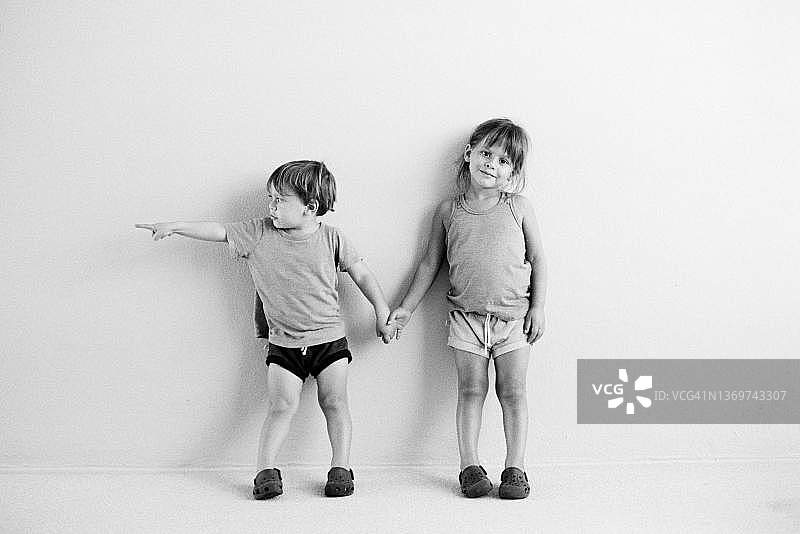 姐弟牵手站在白色墙壁前，妹妹四岁，微笑看着镜头，弟弟两岁，指着画面外的东西图片素材