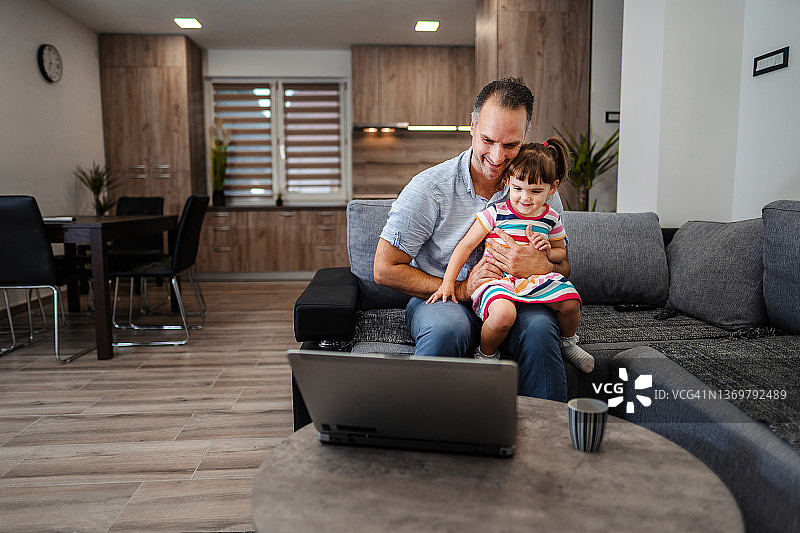 可爱的小女孩和她的父亲在客厅里用笔记本电脑看卡通片图片素材
