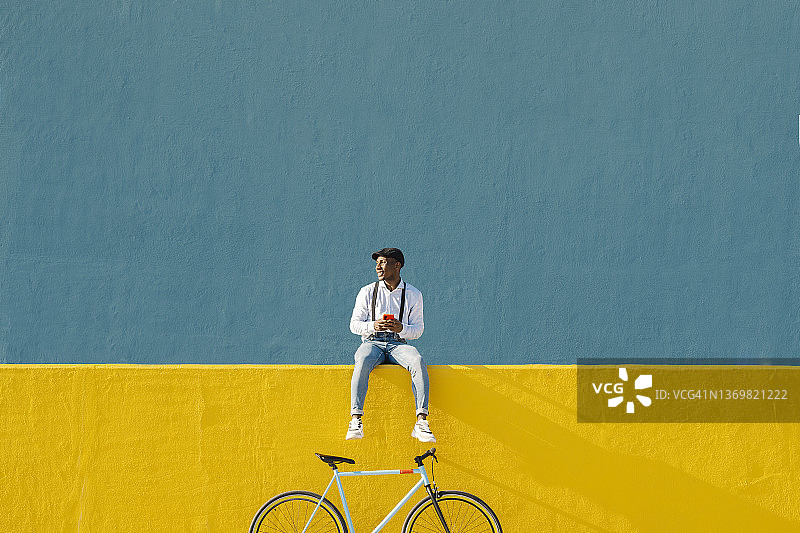 坐在黄色墙边的拿手机和自行车的年轻男人图片素材