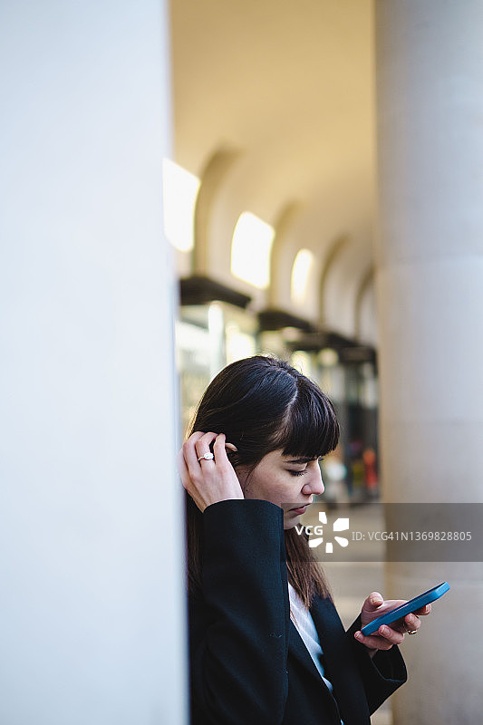 在柱子旁用智能手机，手放在头发上的年轻女人图片素材