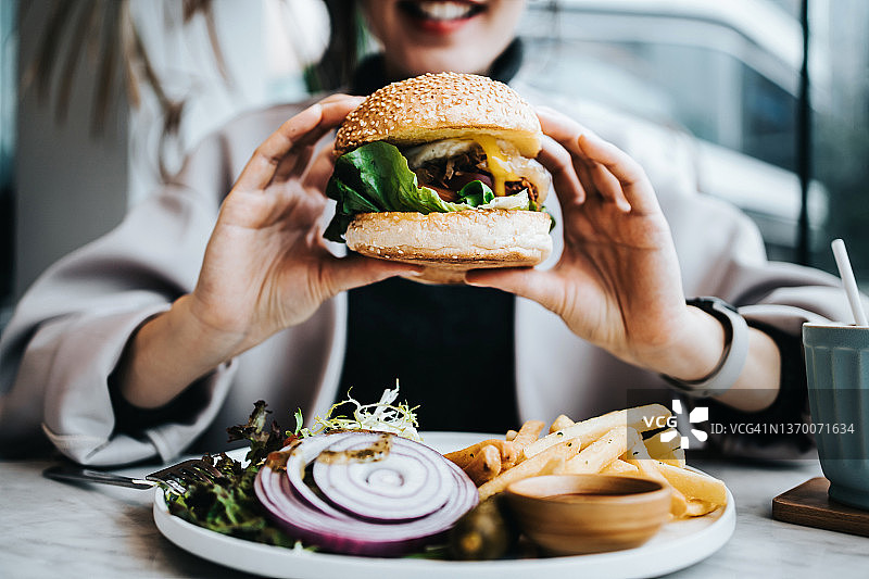 快乐的年轻亚洲女性坐在餐厅的窗边，享受午餐的特写。她正在吃新鲜制作的美味汉堡，配有炸薯条和沙拉。生活方式、人物和食物概念图片素材