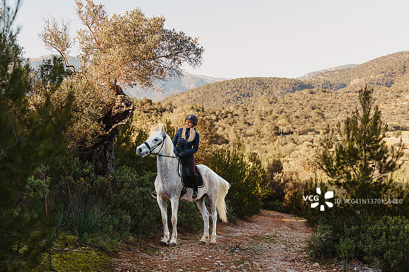 快乐的金发女子在马略卡岛的橄榄树林中骑马图片素材