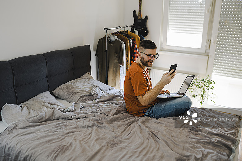 在卧室用手机和笔记本电脑工作的年轻人图片素材