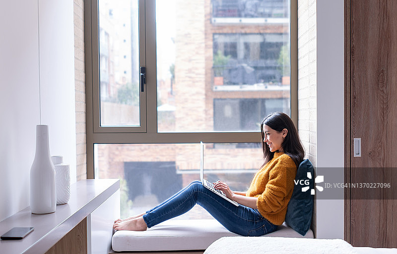 坐在窗边使用笔记本电脑的快乐拉丁美洲年轻女性图片素材