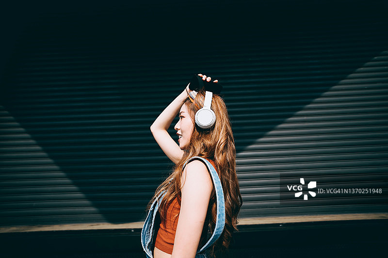 快乐的亚洲年轻女子在户外彩色墙壁和阳光下戴着耳机听音乐跳舞，手持智能手机的侧面轮廓。音乐和生活方式图片素材