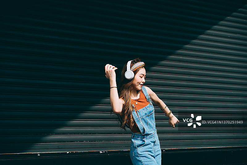 戴着耳机，在彩色墙壁和阳光下跳舞的亚洲年轻女子图片素材