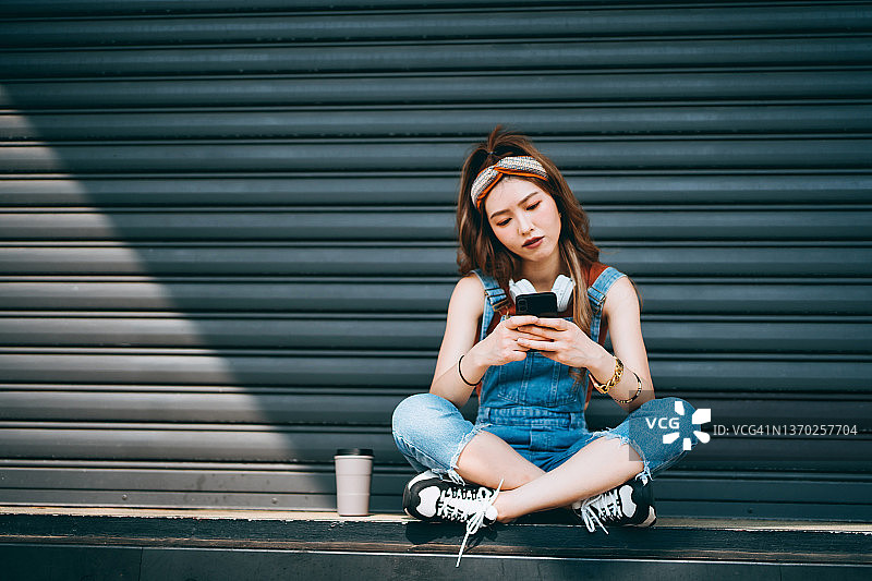 时尚的亚洲年轻女人戴着耳机，在智能手机上滑动，旁边放着一个可重复使用的咖啡杯图片素材