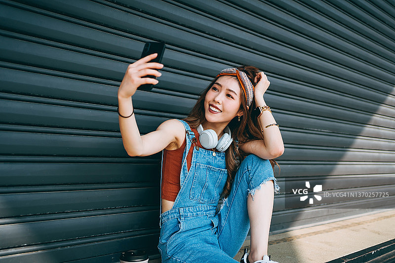 时尚微笑的亚洲年轻女性戴着耳机，坐在卷帘门前，用智能手机自拍。青少年生活方式和科技图片素材