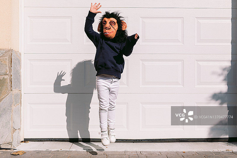 戴乳胶猴子面具的小孩在白色车库门口挥舞手臂跳舞，伪装嘉年华，奇怪有趣的炫酷概念图片素材