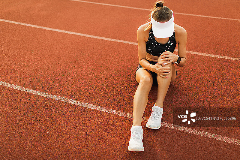 坐在运动跑道上，膝盖受伤并扭伤韧带，感到痛苦的女运动员图片素材