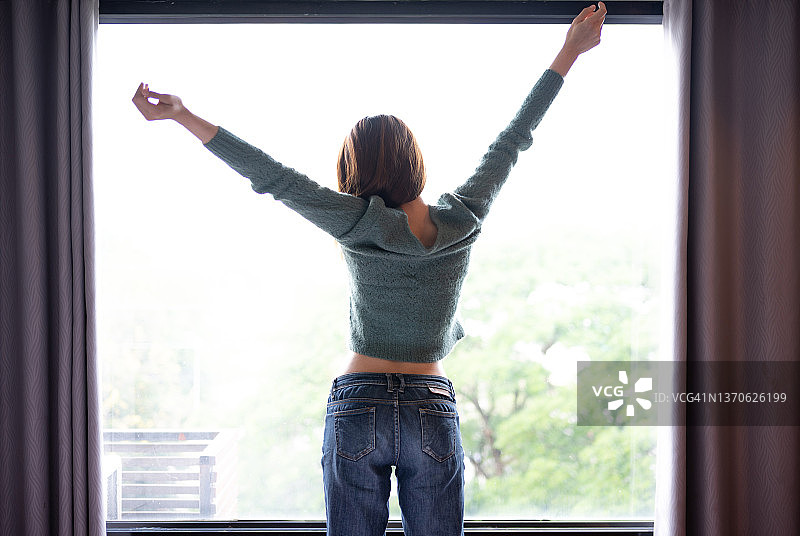 年轻女子站在窗边，伸手伸展，眺望窗外图片素材