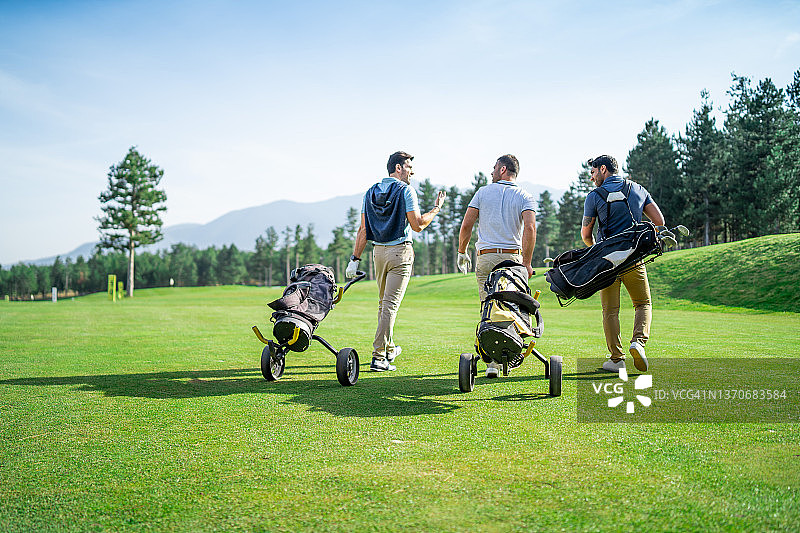 三位男性高尔夫球友在阳光明媚的日子里打高尔夫球的背面，带着高尔夫装备走在高尔夫球场上图片素材