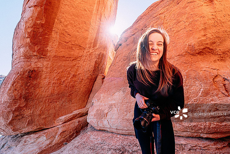 一位快乐的年轻女子站立的广角肖像，她向前倾，笑着，在科罗拉多国家纪念碑参观和徒步旅行时，拿着一台专业的数码单反相机在户外图片素材