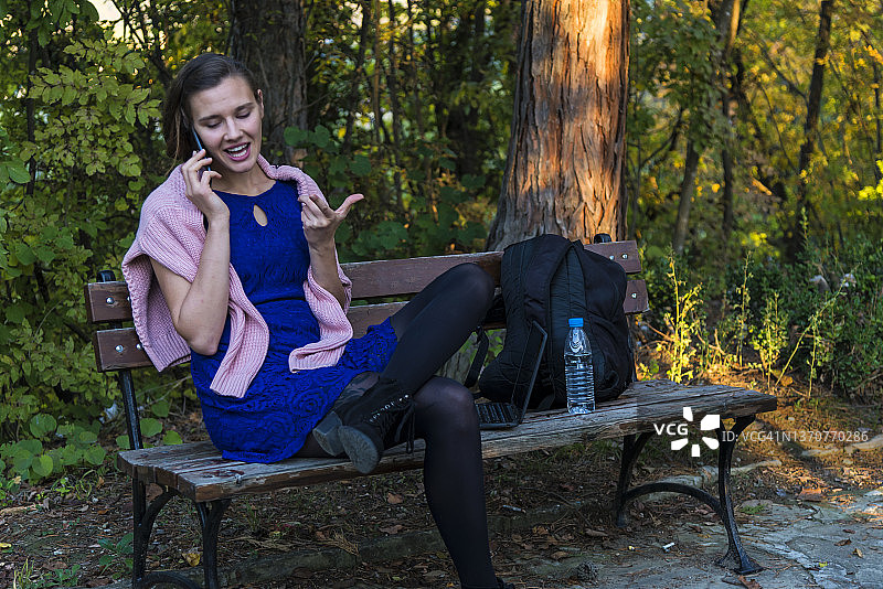 年轻女人坐在公园的椅子上打电话。图片素材
