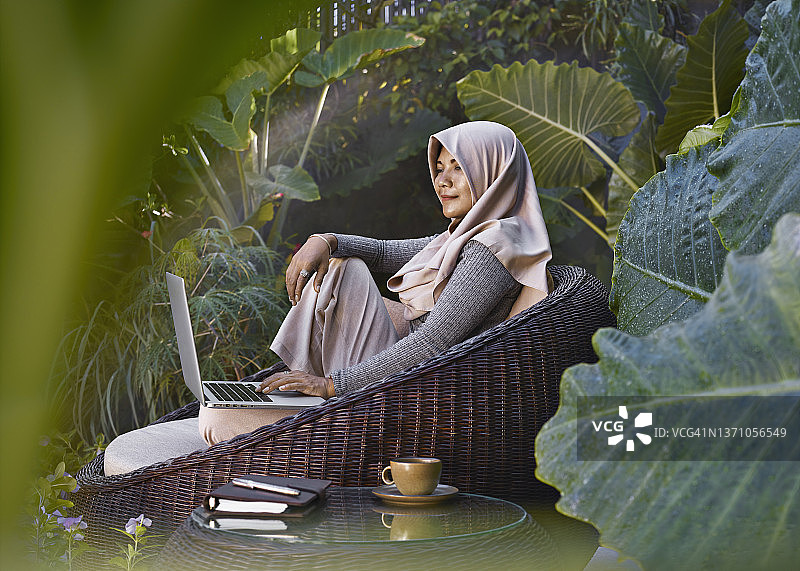亚洲女人戴着头巾，坐在花园家具上在热带植物的环绕下在家工作，使用笔记本电脑图片素材