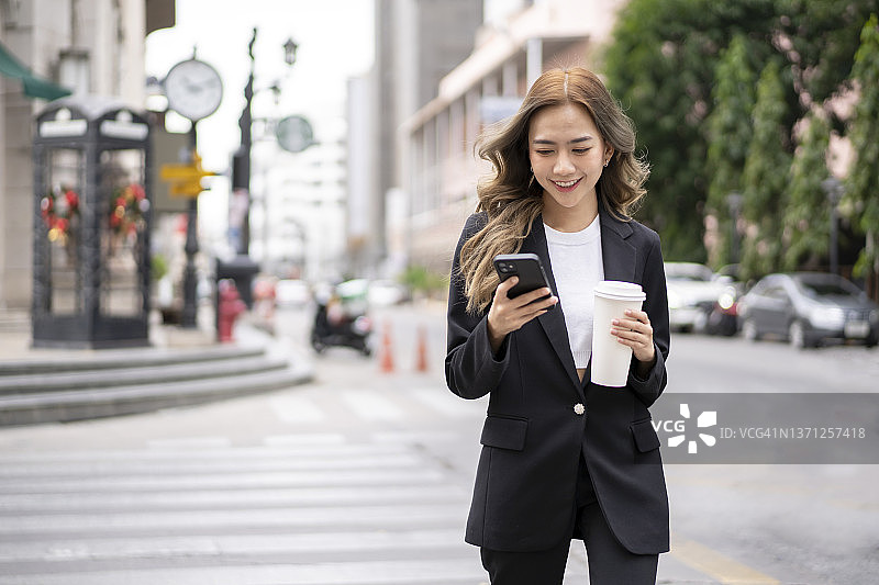 拿着可重复使用马克杯，在城市街道上边走边用智能手机交谈的美丽女商人图片素材