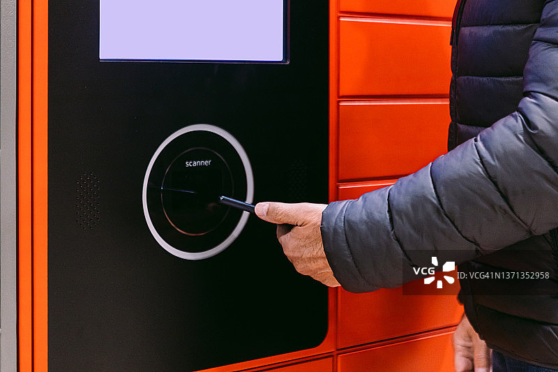 老年男人用手机扫描红色储物柜上的代码以接收包裹的细节：快递、网购与电子商务概念图片素材