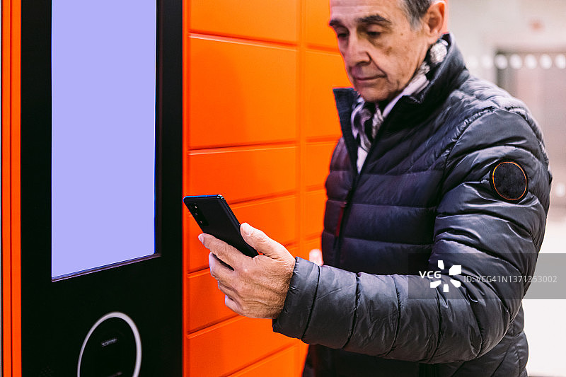 穿着外套的年长男士扫描手机，然后从红色储物柜中取包裹。快递、网上购物、电子商务和包裹的概念图片素材