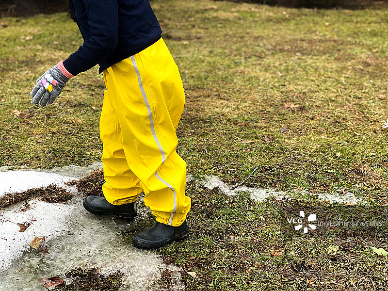 年轻女孩穿着亮黄色雨裤和橡胶靴，在外面玩耍最后的雪图片素材