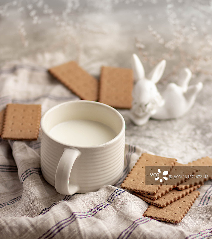 一个白色陶瓷马克杯，里面有牛奶、饼干和一个白色陶瓷兔子，周围环绕着小白花，清淡的早餐图片素材