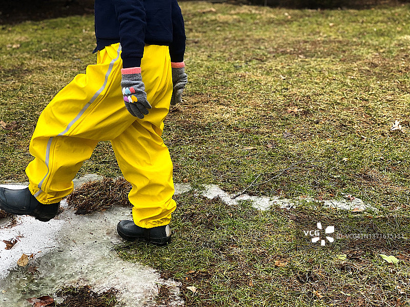 年轻女孩穿着亮黄色雨裤和橡胶靴，在外面玩耍最后的雪图片素材