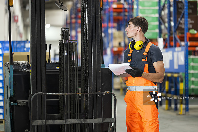 电动叉车预操作清单。男性维修技术员或叉车司机正在检查电池约束装置是否正常工作，并在工厂仓库记录检查表报告。图片素材