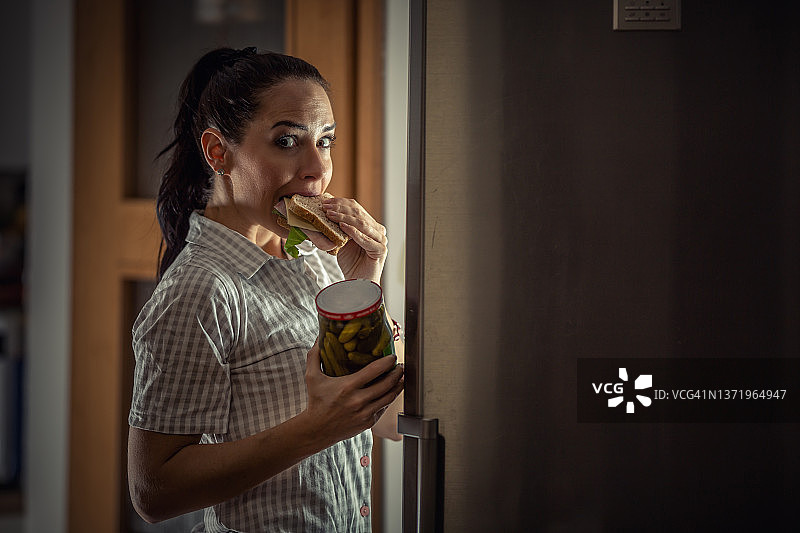 女人晚上翻冰箱，咬着三明治，手里拿着腌黄瓜，面带愧疚图片素材