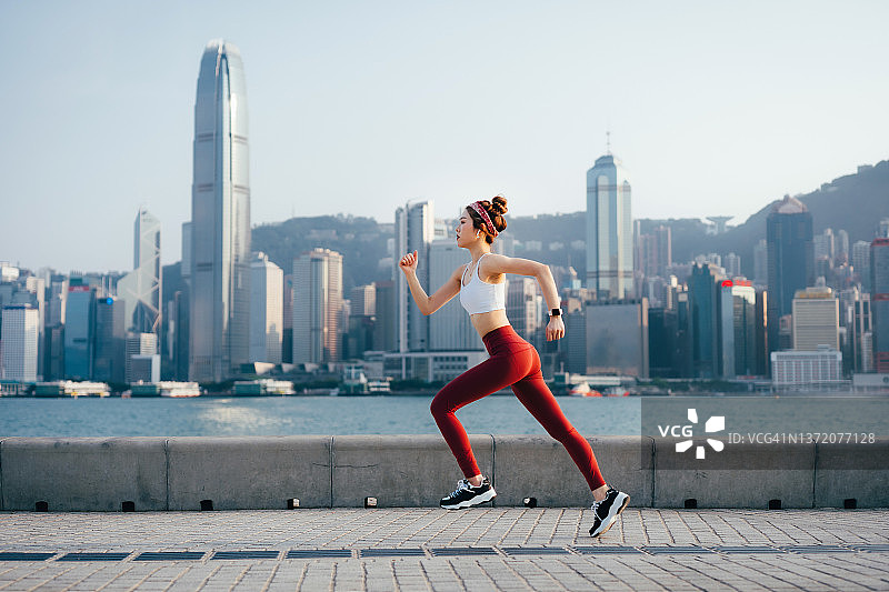 充满活力的年轻亚洲女性戴着耳机听音乐，在城市公园的海滨长廊跑步的侧面轮廓。积极健康的生活方式。户外跑步。健康和健身概念图片素材
