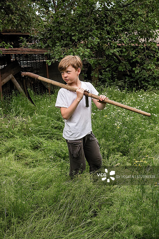 男孩用手镰刀以传统方式在村庄里割草图片素材