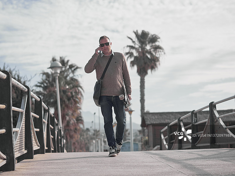 穿着休闲装、拿着笔记本电脑包、胳膊下夹着滑板，边打电话边去上班的商务男士图片素材