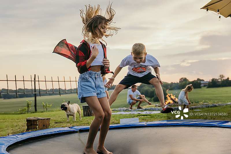 傍晚，女孩和弟弟在蹦床上跳跃，背景是篝火和狗图片素材