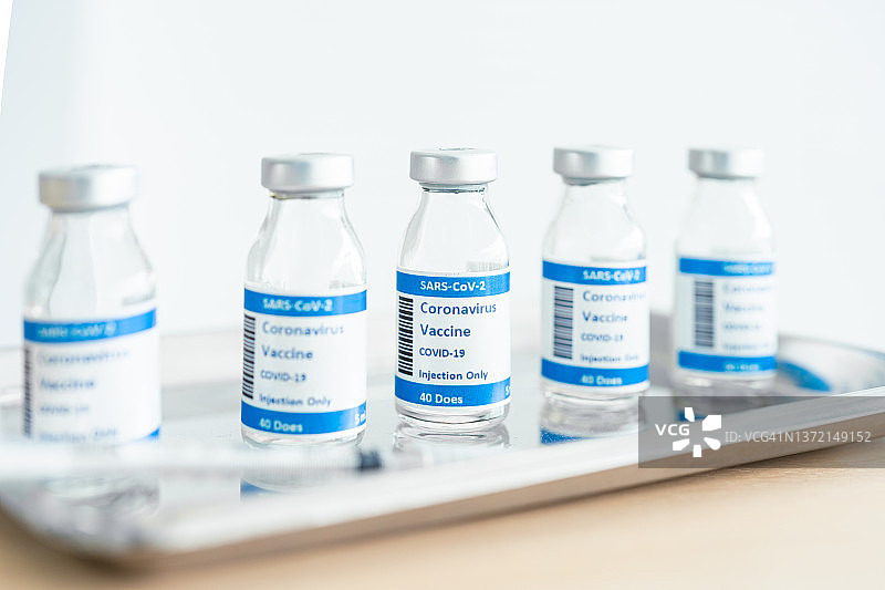 实验台上装有新冠疫苗的小瓶，背景中一位医护专业人员正在整理一盘新冠疫苗小瓶和注射器图片素材