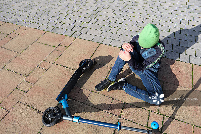 一个青少年站在地上的滑板车旁边，揉着他瘀伤的膝盖。户外。极限运动和青年活动的概念图片素材