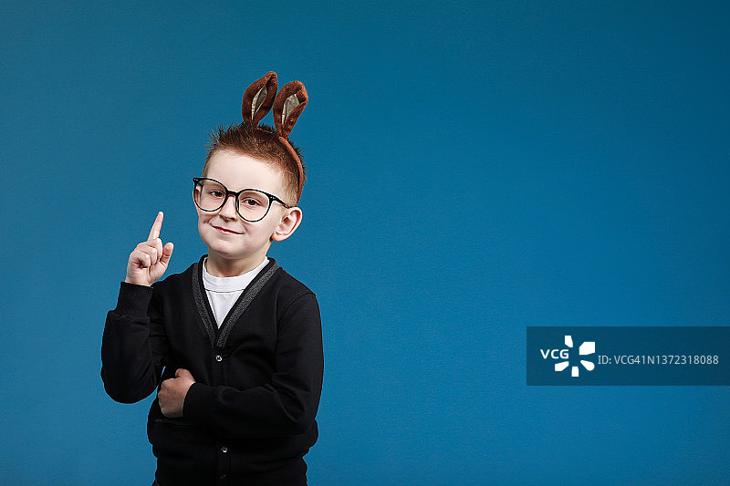 复活节快乐儿童。戴着兔子耳朵的男孩，戴着眼镜，蓝色工作室背景。指向上方。看着镜头微笑。图片素材