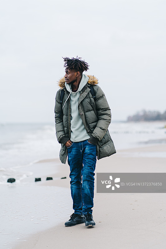 穿着暖和的衣服，背着背包，走在海边丁坝附近的沙滩上，面带微笑的男人，双手插在口袋里图片素材