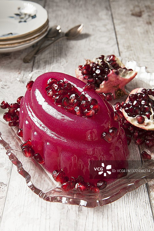 闪光粉色果冻，覆盖着石榴籽，放在复古玻璃盘中，背景是切开的石榴图片素材