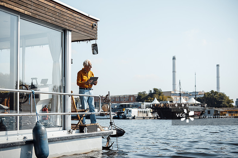 在阳光明媚的日子里，站在船屋栏杆旁手持平板电脑的年长男人图片素材