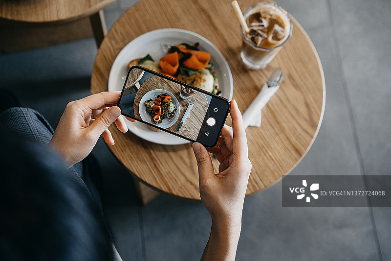 俯视视角：亚洲女人拍摄美味早餐，木桌上放着搭配烟熏三文鱼的班尼迪克蛋。在咖啡馆，一位女性用智能手机拍摄食物。外出就餐生活方式，美食博主，相机先吃文化图片素材