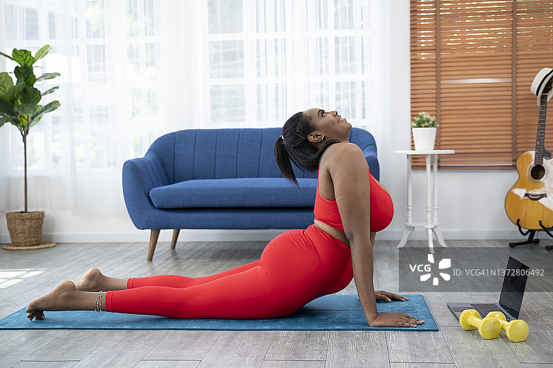 在家锻炼的加大码非裔女士，使用笔记本电脑与在线私人教练进行肘部平板支撑图片素材