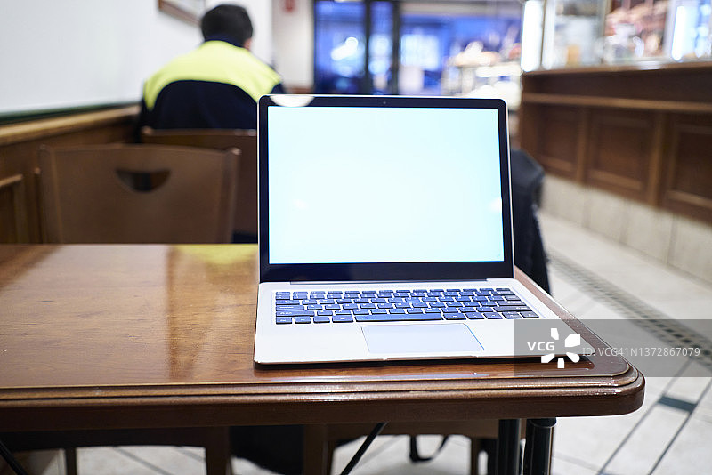 咖啡店木桌上白色屏幕的笔记本电脑的正面特写，背景中坐着一个无法辨认的男人图片素材