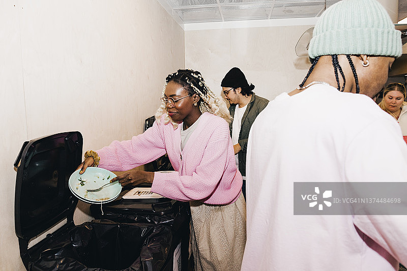 年轻女人在大学宿舍厨房里刮垃圾桶里的剩菜，朋友们在旁边图片素材