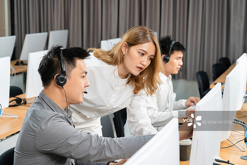 呼叫中心员工使用电脑和耳机，女经理向新同事讲解营销数据库程序，繁忙的客服电话接线员坐在共享办公桌前，协助团队合作概念图片素材