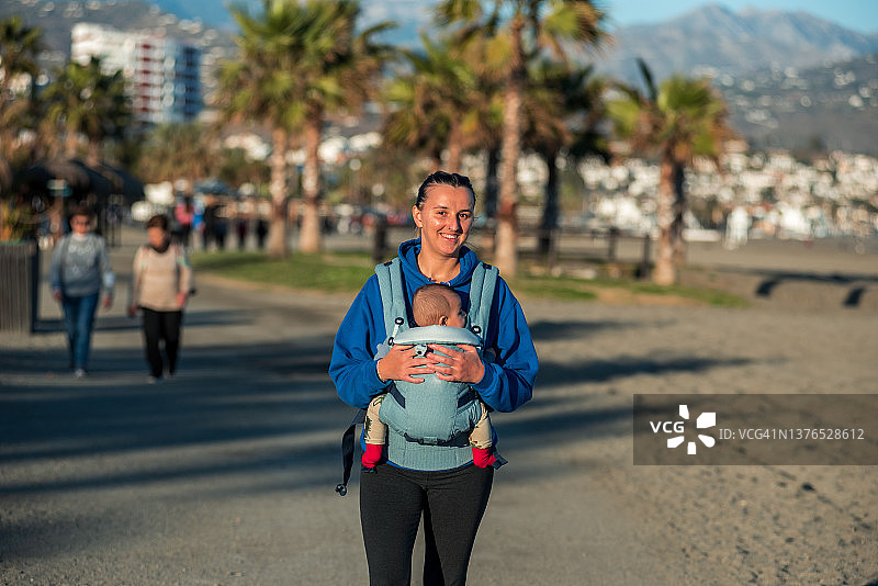 年轻女子用婴儿背带带着宝宝在海滩散步图片素材