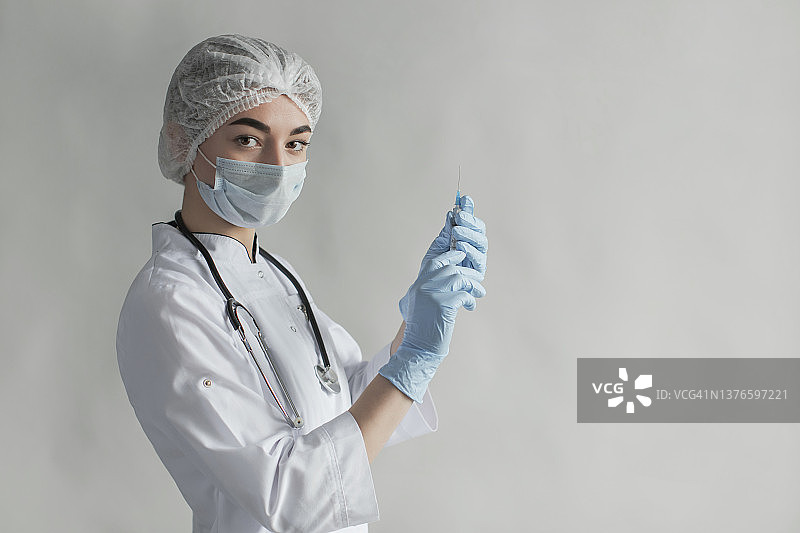 新冠疫苗加强针。带针头的注射器。灰色背景中戴防护口罩和手套的医生肖像。穿专业制服的医疗专家。自信的护士。病毒流行图片素材
