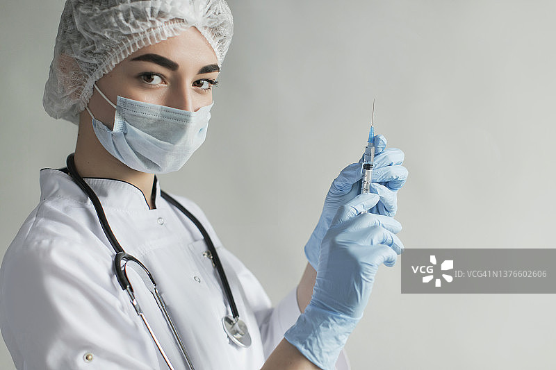 新冠疫苗加强针。带针头的注射器。灰色背景中戴防护口罩和手套的医生肖像。穿专业制服的医疗专家。自信的护士。病毒流行图片素材